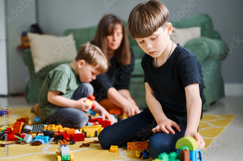Toddler boy playing with construction blocks at home, mother and brother on the background