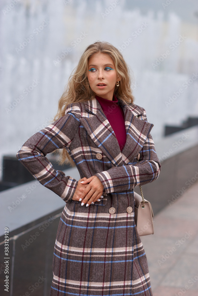 Fototapeta premium Elegant woman walking city street. Fashion fall autumn look. Pretty caucasian blonde stylish girl wearing blue jeans, wine red pullover and squared wool coat. Female with makeup and wavy blonde hair