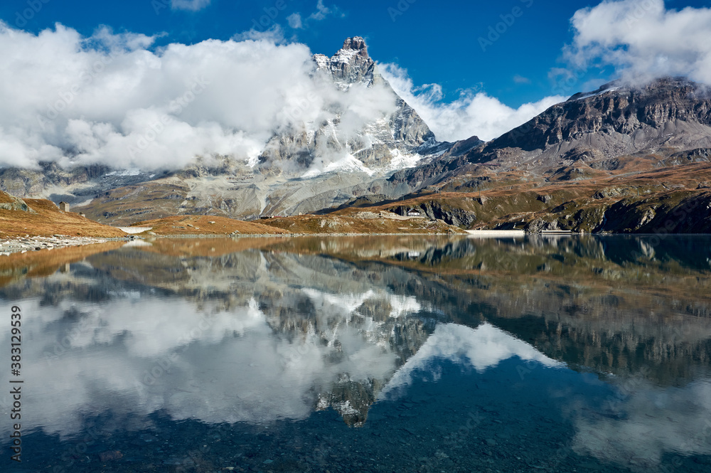 Monte Cervino in floating clouds with reflection in the blue water of a mountain lake against a blue sky