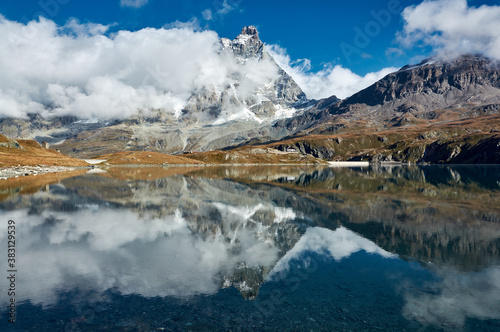 Monte Cervino in floating clouds with reflection in the blue water of a mountain lake against a blue sky