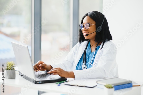 Smiling african american female doctor gp wears white medical coat using laptop computer at workplace gives remote online consultation, working on pc, consulting patient in internet telemedicine chat.