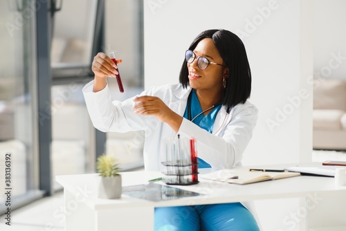 Young beautiful African American girl doctor in a white coat with a stethoscope. sitting at a table with reagent flasks on white background. Female lab assistant studying blood sample for analysis