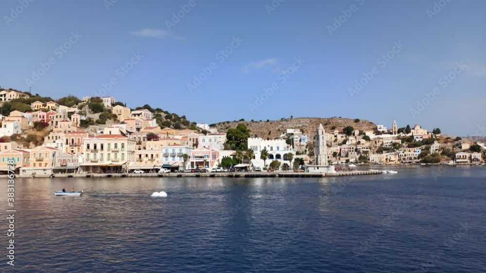 Rhodes, Greece: Pan shot of symi island with colorful houses on rusty mountain situated in mediterranean sea against clear blue sky Rhodes, Greece: Pan shot of symi island with colorful houses on rusty mountain situated in mediterranean sea against clear blue sky