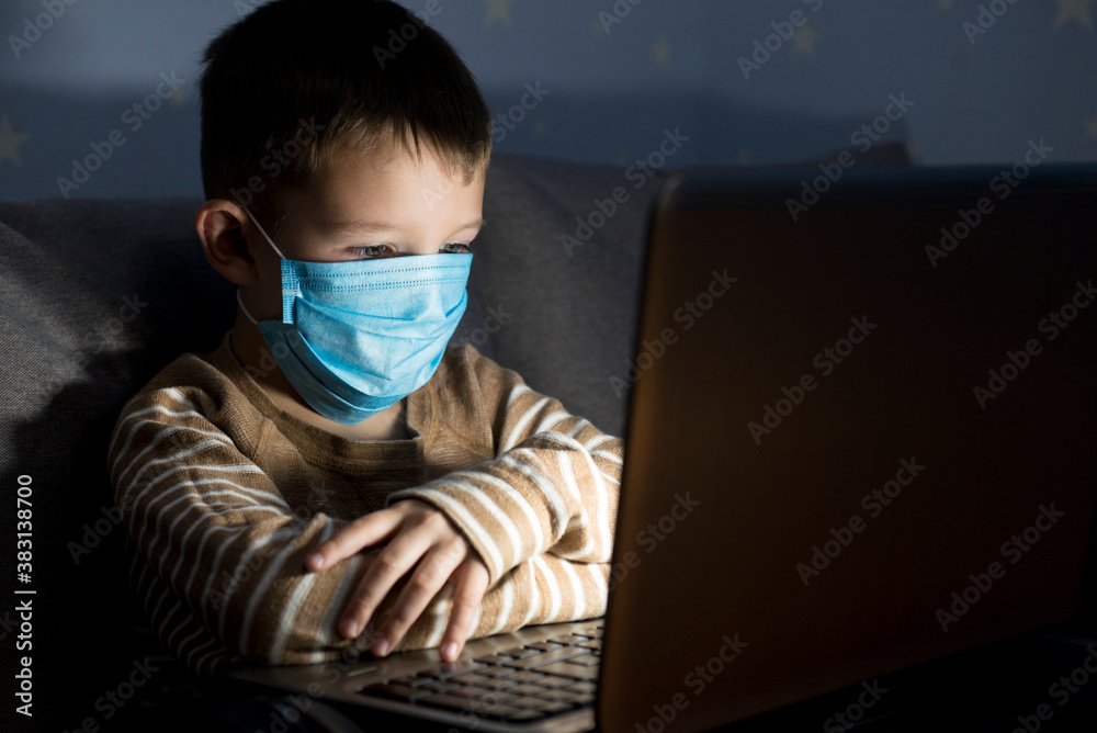 Child in medical mask is looking at notebook and getting the new information in the evening, online education during quarantine Child in medical mask is looking at notebook and getting the new information in the evening, online education during quarantine