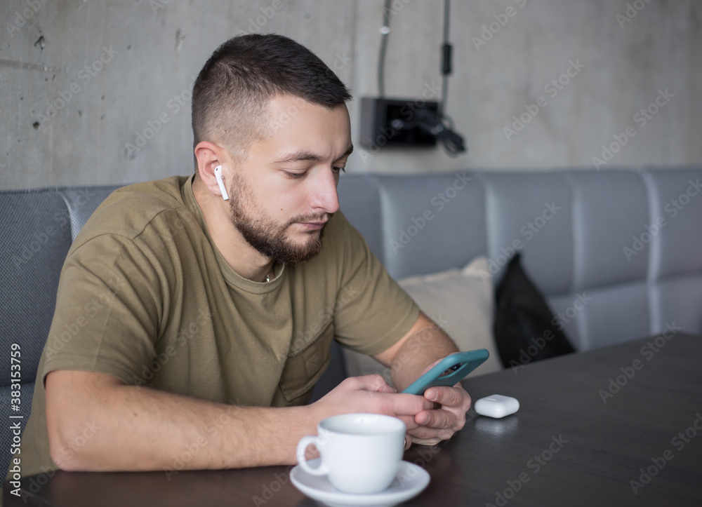 Young man in casual clothes listening to music in wireless earphones while using smartphone in cafe.