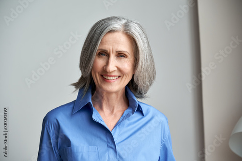 Smiling sophisticated mature grey-haired woman standing on grey wall background at home. Happy elegant middle aged old lady professional businesswoman entrepreneur posing in office, headshot portrait. photo
