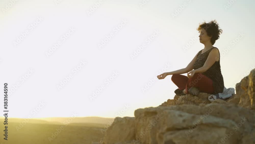 Young woman relaxing practicing meditation on a desert mountain top at sunset. Self examination, concentration and deep thought on a cliff with epic and beautiful colors. Sits quiet and peaceful. 