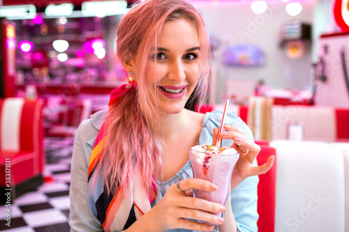 Cute pretty sensual woman with trendy pink hairs enjoy her sweet strawberry milk shake, smiling and looking at camera, fashionable vintage pastel outfit.