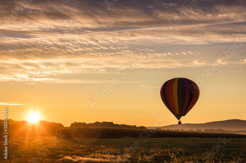 Hot Air Balloon in colorful rainbow stripes begins ascent over farm field as sun rises golden sky