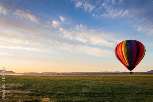 Hot Air Balloon in colorful rainbow stripes begins ascent over farm field as sun rises blue cloudy sky © rabbitti