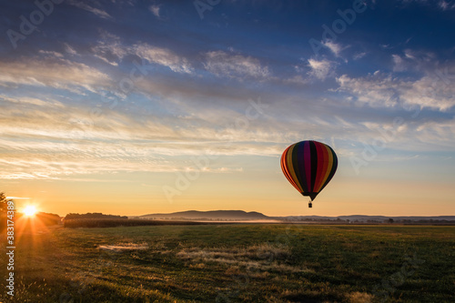 Hot Air Balloon in colorful rainbow stripes begins ascent over farm field as sun rises blue cloudy sky