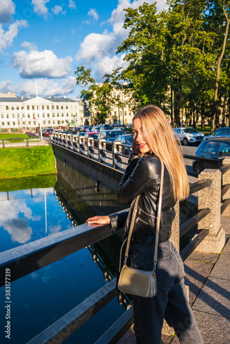 A beautiful girl in a half-rotation looks to the side. A girl in a leather jacket against the background of a white building holds her face with her hands. Long hair in a white European girl.