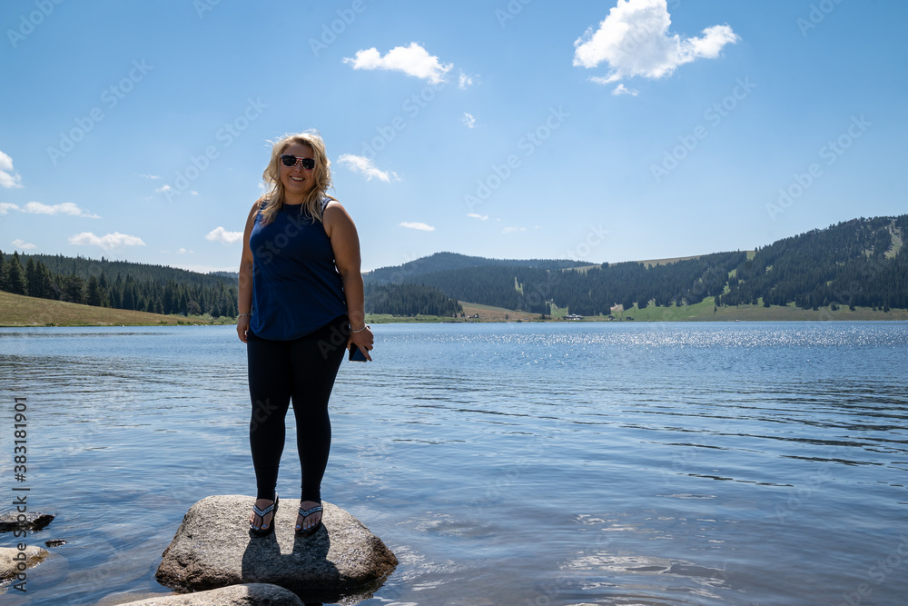 Happy smiling blonde woman stands on a rock in Meadowlark Lake in Wyoming, holding a phone