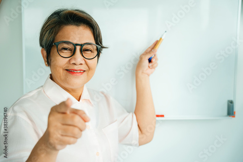 Elderly senior old teacher woman standing near white board and pointing at camera.