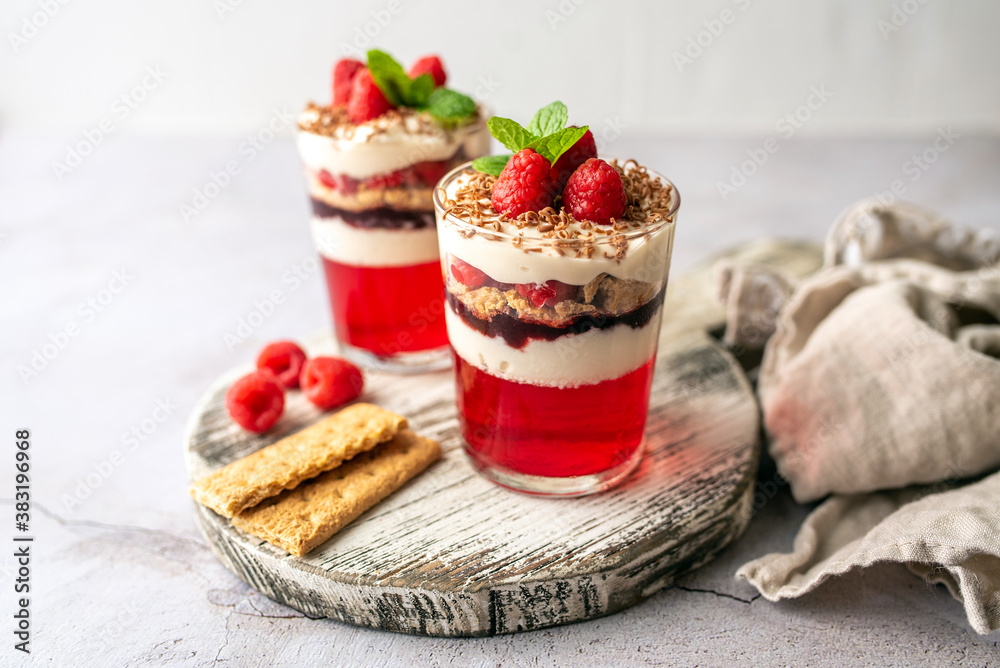 Dessert in a glass cup, with Jello covered in yogurt and topped with fresh raspberries. Mini dessert on light concrete background. Fruit parfait with jello and jelly.  - obrazy, fototapety, plakaty 