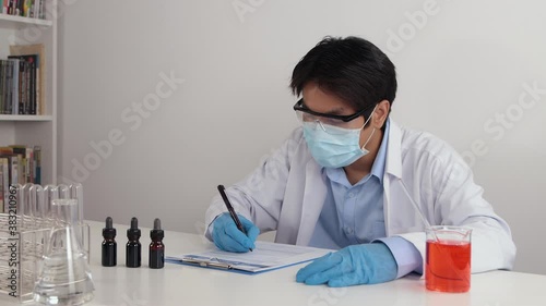 Asian Doctor or Scientist Wear Face Mask and White Coat or Gown Writing Experiment Report in Lab in Corona Virus Disease or Covid 19 Situation