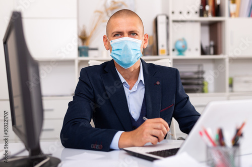 Portrait of young man in medical mask sitting at desk with laptop and working in office