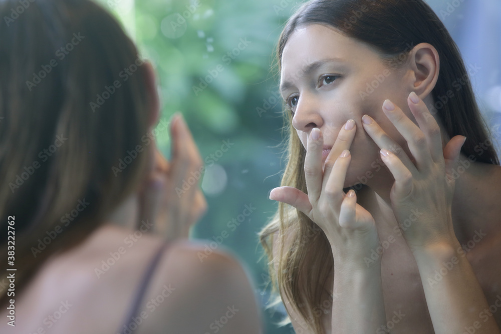 Portrait of a beautiful young woman standing in the bathroom and examining her face in the mirror, problematic acne-prone skin concept Portrait of a beautiful young woman standing in the bathroom and examining her face in the mirror, problematic acne-prone skin concept