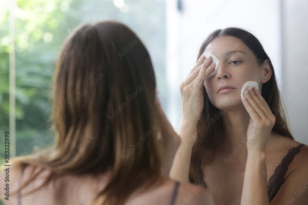 Portrait of a beautiful young woman standing in the bathroom and cleaning her face with cotton pads in front of the mirror, removing makeup, beauty and spa concept. Portrait of a beautiful young woman standing in the bathroom and cleaning her face with cotton pads in front of the mirror, removing makeup, beauty and spa concept.