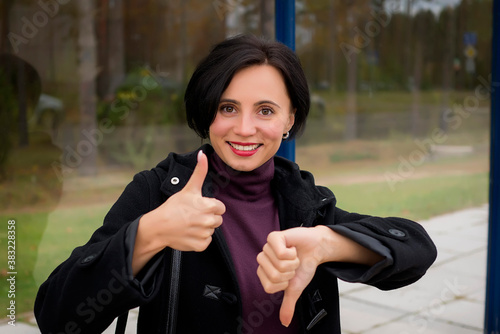 A young beautiful woman in a black coat laughs happily and shows thumbs up and down.
