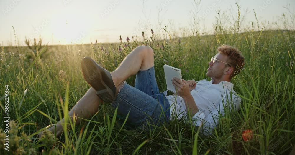 Young male poet lying on green grass and writing in notepad while creating poem in meadow at sunset Young male poet lying on green grass and writing in notepad while creating poem in meadow at sunset