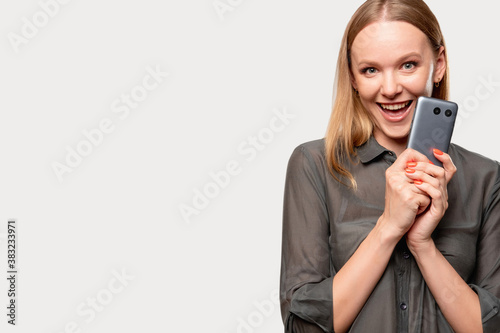Modern device. Female user. Technology addiction. Excited amused woman in gray shirt holding new mobile phone smiling isolated on light neutral empty space background.