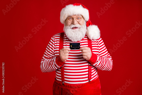 Photo of retired grandpa grey beard hold debit card prepare cashless online shopping sale wear santa x-mas costume suspenders spectacles striped shirt cap isolated red color background © deagreez