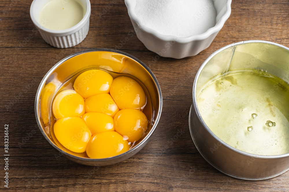 Raw ingredients in containers for baking, making a cake, or other desserts. Raw separated egg yolks, egg white, white sugar, cream. On a wood table top Raw ingredients in containers for baking, making a cake, or other desserts. Raw separated egg yolks, egg white, white sugar, cream. On a wood table top