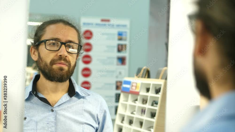 Bearded man is choosing eyesight glasses at optics store. Male is trying eyeglasses while looking at mirror. Bearded man is choosing eyesight glasses at optics store. Male is trying eyeglasses while looking at mirror.