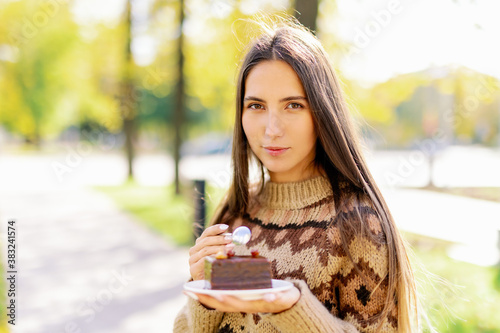 Smiling caucasian female model eating fancy slice cake in outdoor on background of green park, enjoying gentle chocolate dessert and coffee , girl eat sweet cake