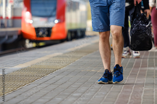 Legs of a man in shorts and sneakers walking along the platform of a railway station against the background of an arriving train on a sunny summer day. In the hands of a small bag. Travel light.