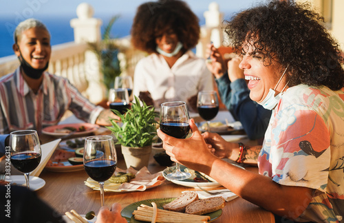 Young multiracial people eating and drinking red wine while wearing protective masks - Happy friends having fun doing dinner at restaurant patio - Social distance concept - Focus on right girl face