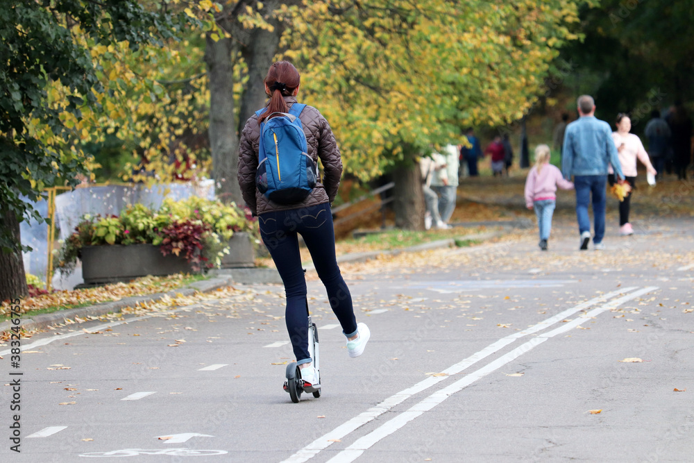 Woman rides an electric scooter on a city street, rear view. Riding e-scooter in autumn