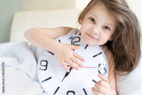 Little sleeping girl with big, huge clock in hands. Early morning wake up before kindergaten,school. White pillow, blanket in bed. Sleepy child. Sweet dreams. Bedtime. Correct mode of children's rest photo