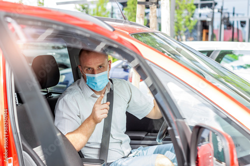 Handsome businessman,looking at camera,wearing protective face mask while sitting in a car.Confident young businessman wearing blue face mask looking at camera while sitting in the car during the day.