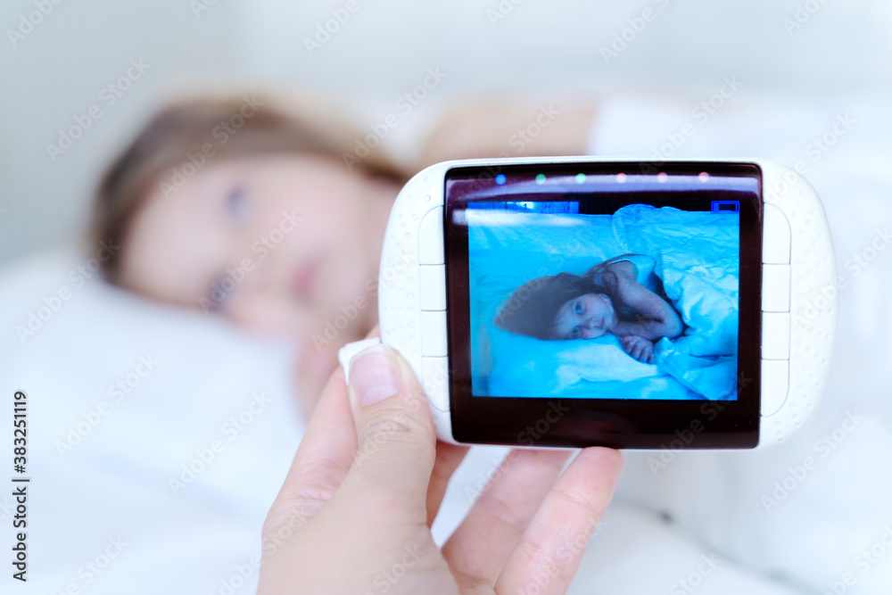 Mom looks at the camera for a little sleeping girl, daughter through the radio, video baby monitor. Protection, care for the safety of the child. Remote parent control. Modern technologies