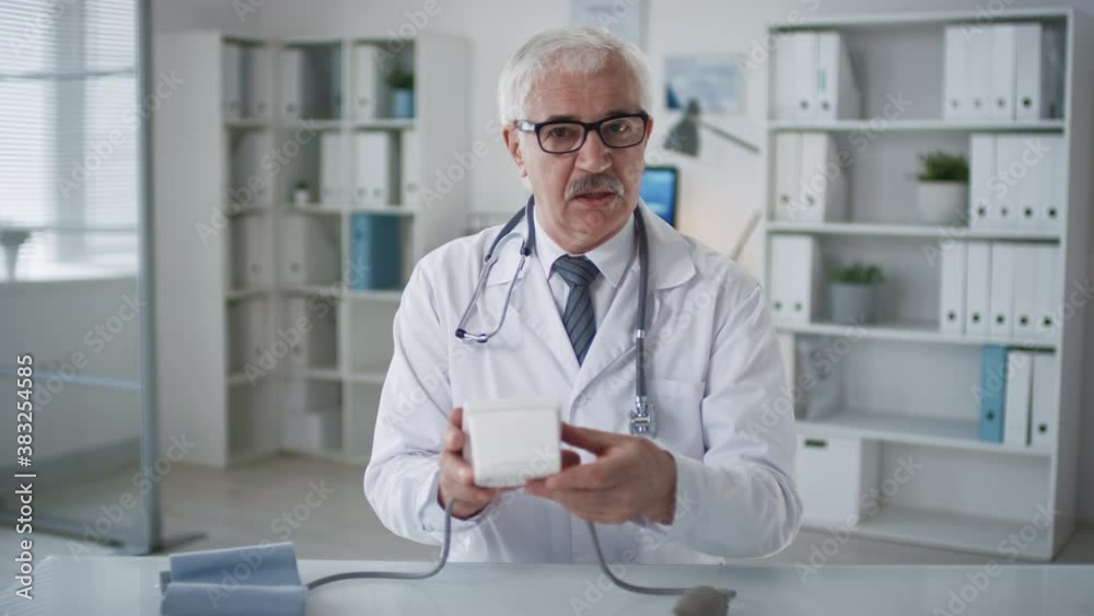 Professional therapist wearing eyeglasses and white coat sitting at desk in his office demonstrating how to use blood pressure monitor on camera Professional therapist wearing eyeglasses and white coat sitting at desk in his office demonstrating how to use blood pressure monitor on camera