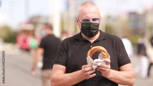 Video, an emotional adult man in a black shirt and sunglasses eats a simit bun on a city street. People are walking next to the man and a tram is passing by. On a sunny day in summer. photo