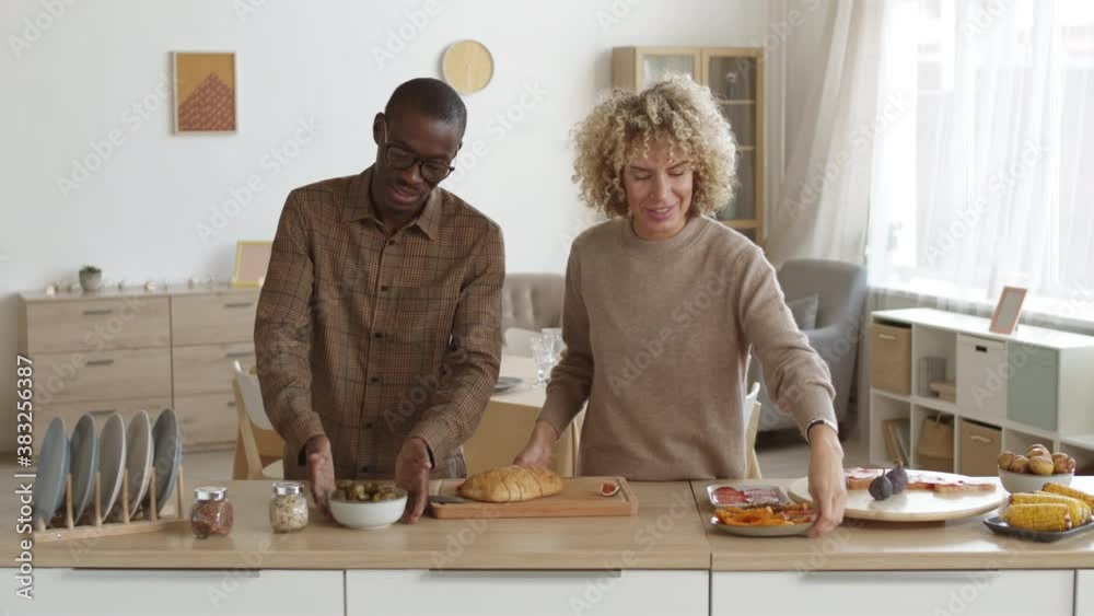 Medium shot of curly-haired Caucasian woman and African-American man standing at kitchen worktop, chatting and cooking in front of camera Medium shot of curly-haired Caucasian woman and African-American man standing at kitchen worktop, chatting and cooking in front of camera