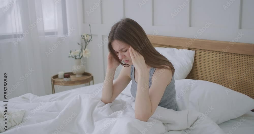 Women sitting on bed holding her head. She has a painful headache. The woman experiences a headache, rubs her temples and frowns in pain. Seasonal disease, Caucasian girl in the bedroom on sick leave Women sitting on bed holding her head. She has a painful headache. The woman experiences a headache, rubs her temples and frowns in pain. Seasonal disease, Caucasian girl in the bedroom on sick leave