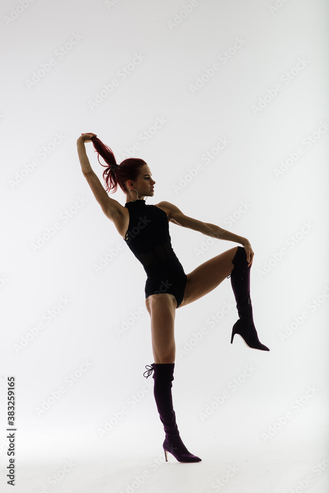 Obraz premium Young beautiful flexible female in black jumpsuit and high heels is posing in a dance studio.