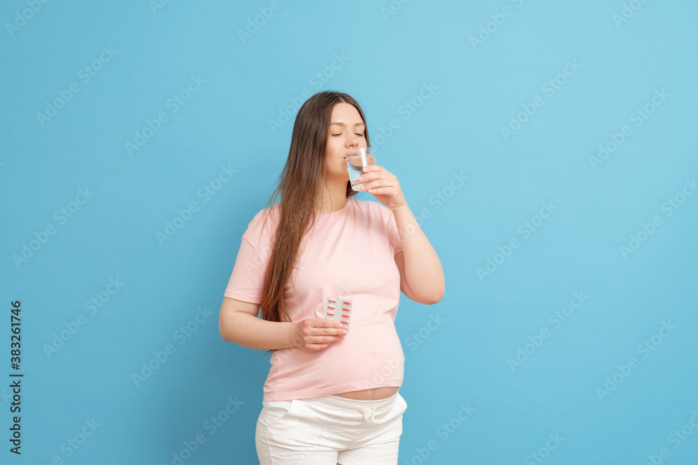 young beautiful pregnant woman on a blue background holds pills in her hand and a glass of water, woman drinking pills young beautiful pregnant woman on a blue background holds pills in her hand and a glass of water, woman drinking pills
