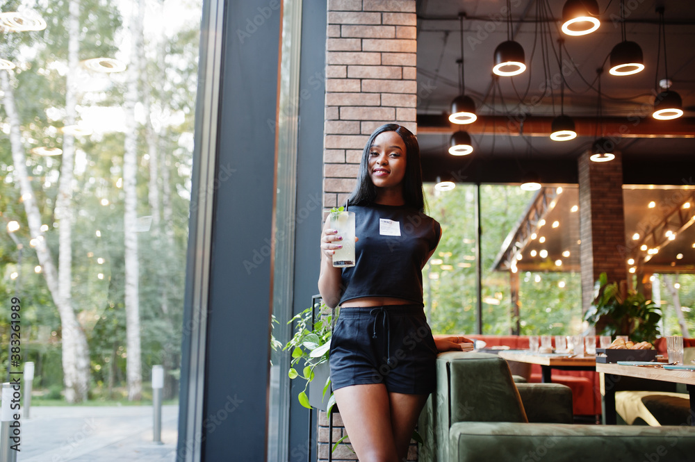 Obraz premium Fashionable feminist african american woman wear in black t-shirt and shorts, posed at restaurant with lemonade glass.