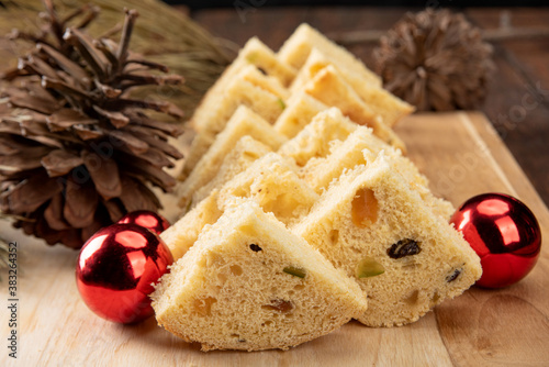 Panettone slices on a polished wood, pine cones and christmas balls on rustic wood, black background, selective focus.