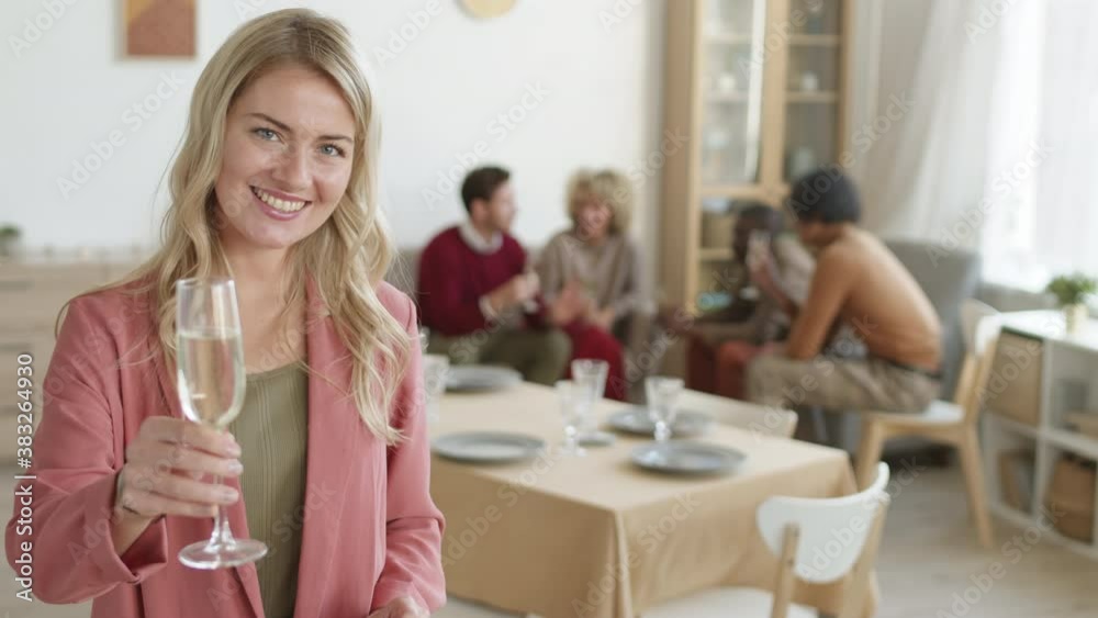 Medium shot of young Caucasian woman standing at kitchen table with champagne glass in hands, looking at camera and making a toast while her friends chatting on background
