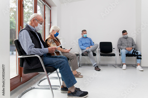 Senior couple with face masks sitting in a waiting room of a hospital together with a young and mature man