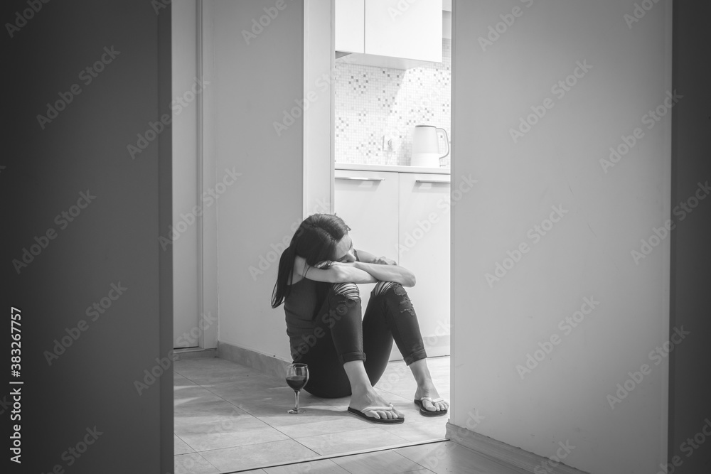 Young caucasian woman sits  on the kitchen doorway sad and lonely with face on her crossed hands and glass of wine on the ground