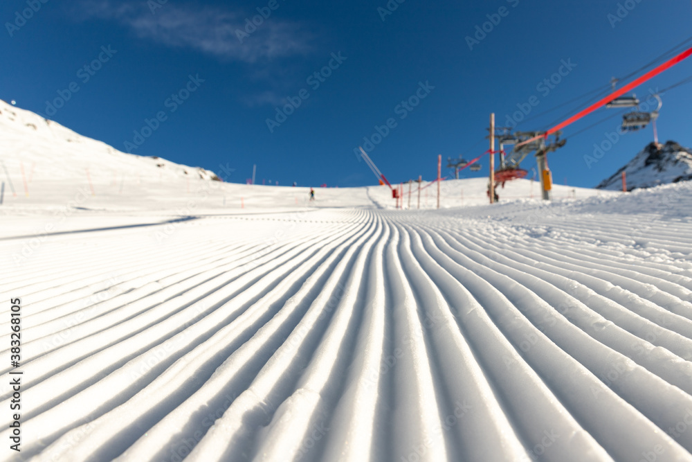 Close-up straight line rows of freshly prepared groomed ski slope piste with bright shining sun and clear blue sky background. Snowcapped mountain downhill landscape at europe winter skiing resort - obrazy, fototapety, plakaty 
