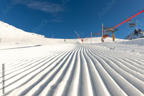 Close-up straight line rows of freshly prepared groomed ski slope piste with bright shining sun and clear blue sky background. Snowcapped mountain downhill landscape at europe winter skiing resort