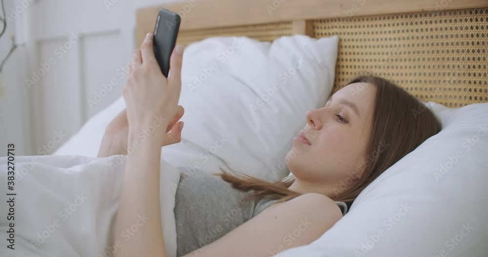 Face Close-up on woman typing a message on a mobile phone using 2 hands while comfortably lying on a bed in a hotel room. woman using smart phone and lying on the bed in Morning after waking up Face Close-up on woman typing a message on a mobile phone using 2 hands while comfortably lying on a bed in a hotel room. woman using smart phone and lying on the bed in Morning after waking up